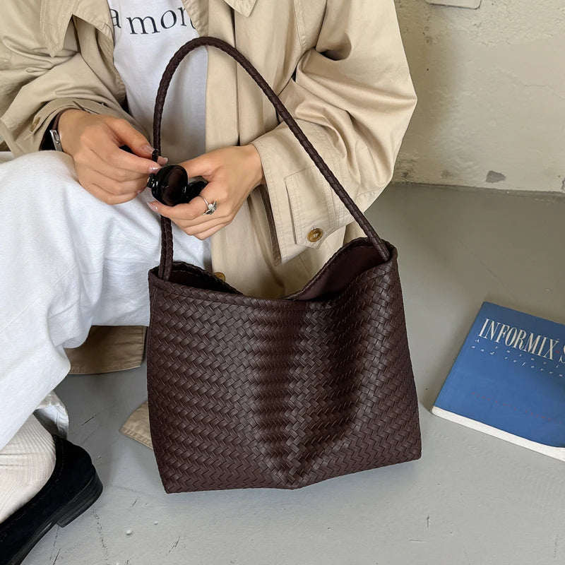 A dark brown woven PU bucket shoulder bag is held by a person wearing a beige jacket and white pants.
