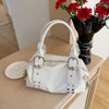 A white PU leather shoulder bag with silver rivets rests on a wooden surface next to a vase of red flowers.