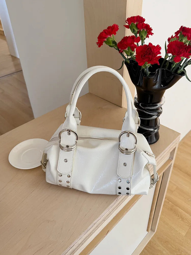 A white PU leather shoulder bag with silver rivets rests on a wooden surface next to a vase of red flowers.