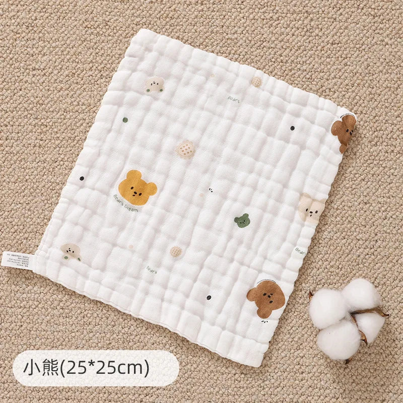 A white quilted muslin baby burp cloth towel with small animal embroidery rests on a beige fabric background.