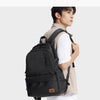 A young man wearing a beige jacket and white shirt carrying a black Oxford school backpack.