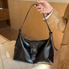 A woman holds a black PU leather underarm bag with a long strap against a light wooden background.