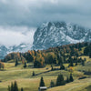 Dramatische Landschaft mit zerklüfteten Bergen, sanften grünen Feldern und dunklen Wolken unter einem stimmungsvollen Himmel.