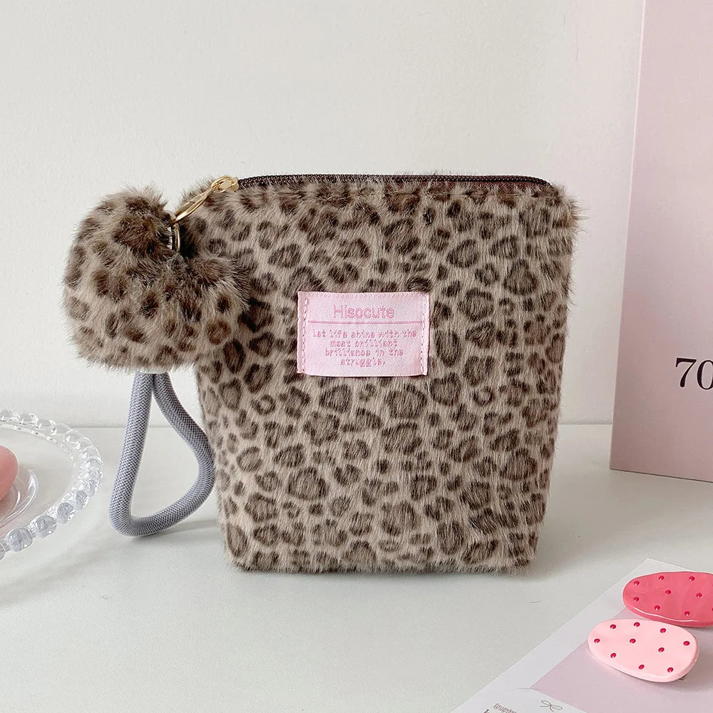 A brown leopard print makeup bag with a fluffy pom-pom and gray strap is displayed on a white surface.
