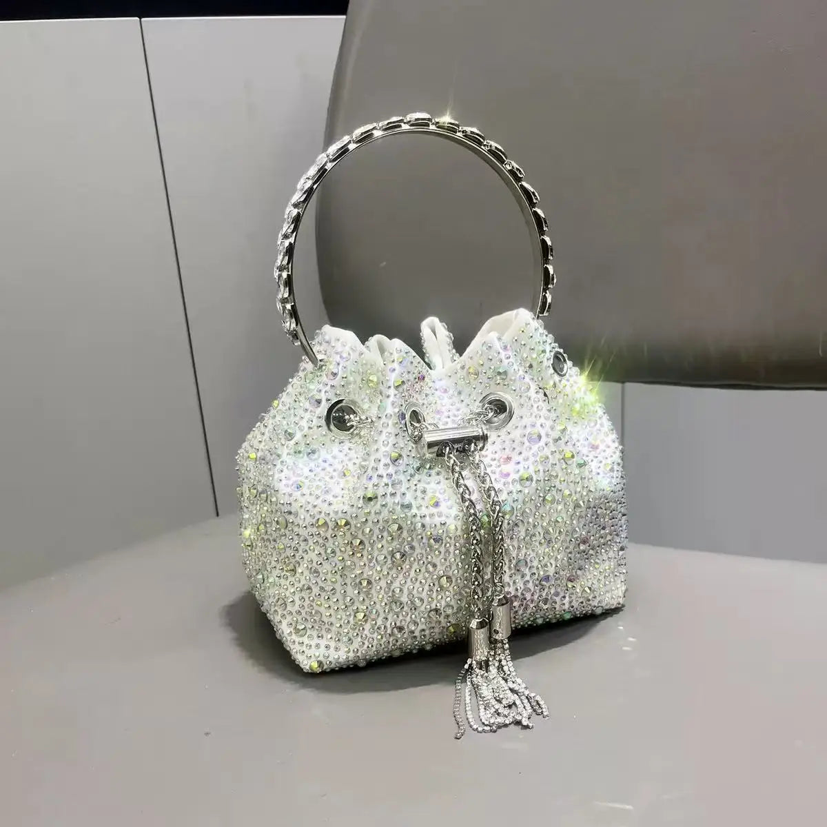 A white crystal-embellished evening clutch with a silver chain strap is displayed on a gray surface.