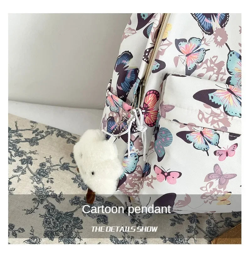A white backpack with a butterfly pattern and a fluffy white charm hanging from the zipper.