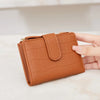A brown PU leather bifold wallet with a zipper closure is held in a hand on a light surface.
