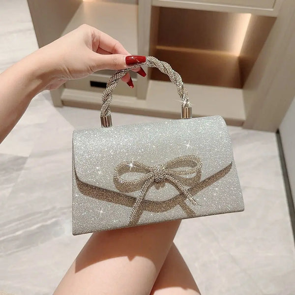 Une main tient un sac à main soirée en nœud pailleté argenté avec une chaîne dorée contre un fond clair.