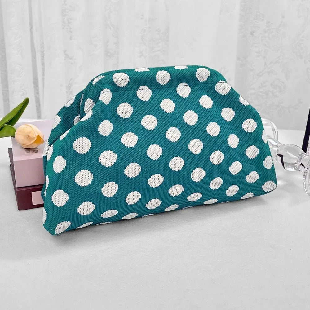 A teal polka dot clutch bag is displayed on a white surface next to a clear glass.