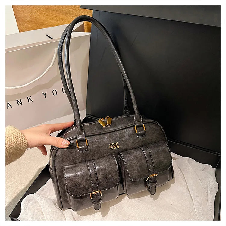 Un bolso de cuero sintético gris oscuro retro con herrajes dorados es sostenido por una mano contra una caja negra.