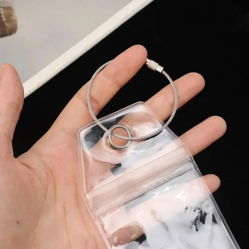 A hand holds a clear luggage tag with a steel ring and a leather cord loop.