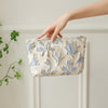 A hand holds a light blue floral canvas makeup bag against a white paneled wall.
