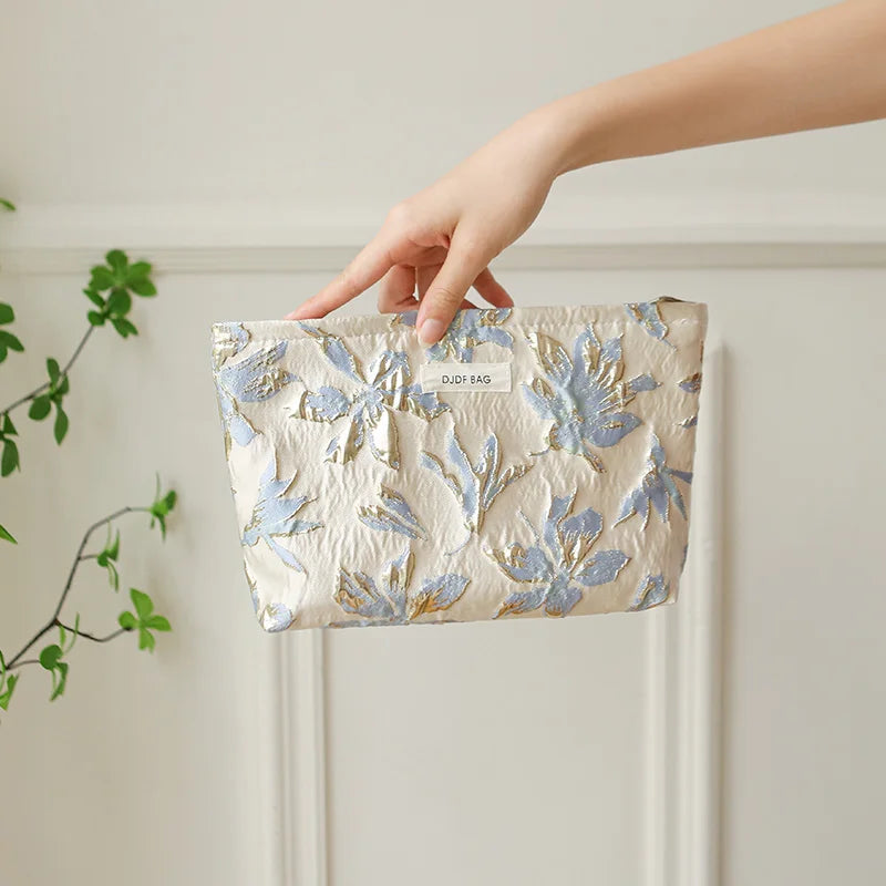 A hand holds a light blue floral canvas makeup bag against a white paneled wall.