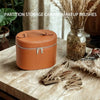 A round, tan leather travel makeup organizer with a zipper closure sits on a wooden surface next to dried decorative grasses.