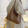 A person wearing a beige trench coat is holding a brown PU leather slouchy hobo shoulder bag.