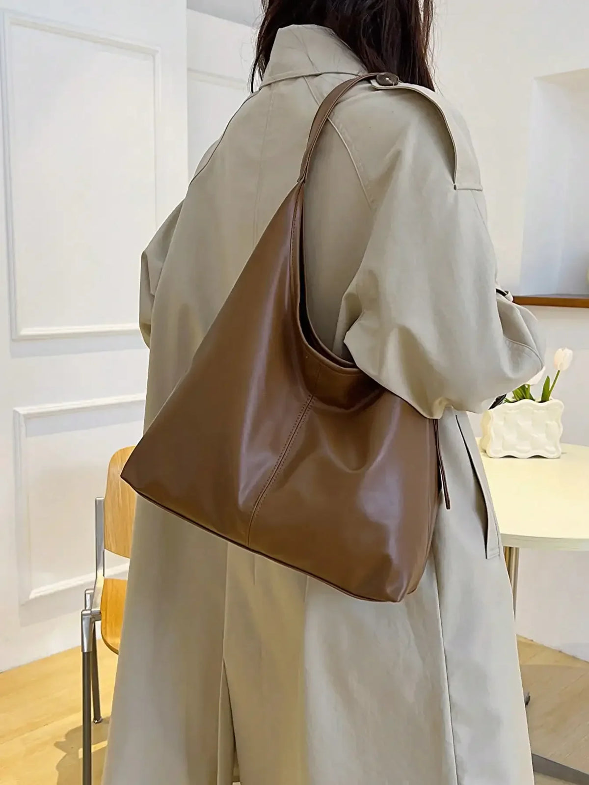 A person wearing a beige trench coat is holding a brown PU leather slouchy hobo shoulder bag.