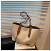 A beige canvas shoulder tote bag with brown leather accents displayed in front of horizontal blinds.
