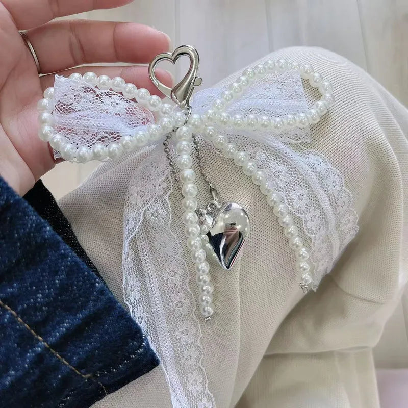 A white lace bow keychain with pearl strands and a silver heart pendant is held in a hand.