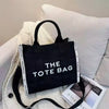A black tote bag with white text is displayed on a white knitted fabric against a light gray background.