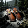 A brown leather knife sheath with a belt loop and decorative stitching rests on a dark stone next to a tactical glove and fab