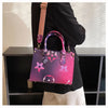 A woman holds a pink and purple patterned ladies' shoulder tote bag with a tan strap against a dark background.
