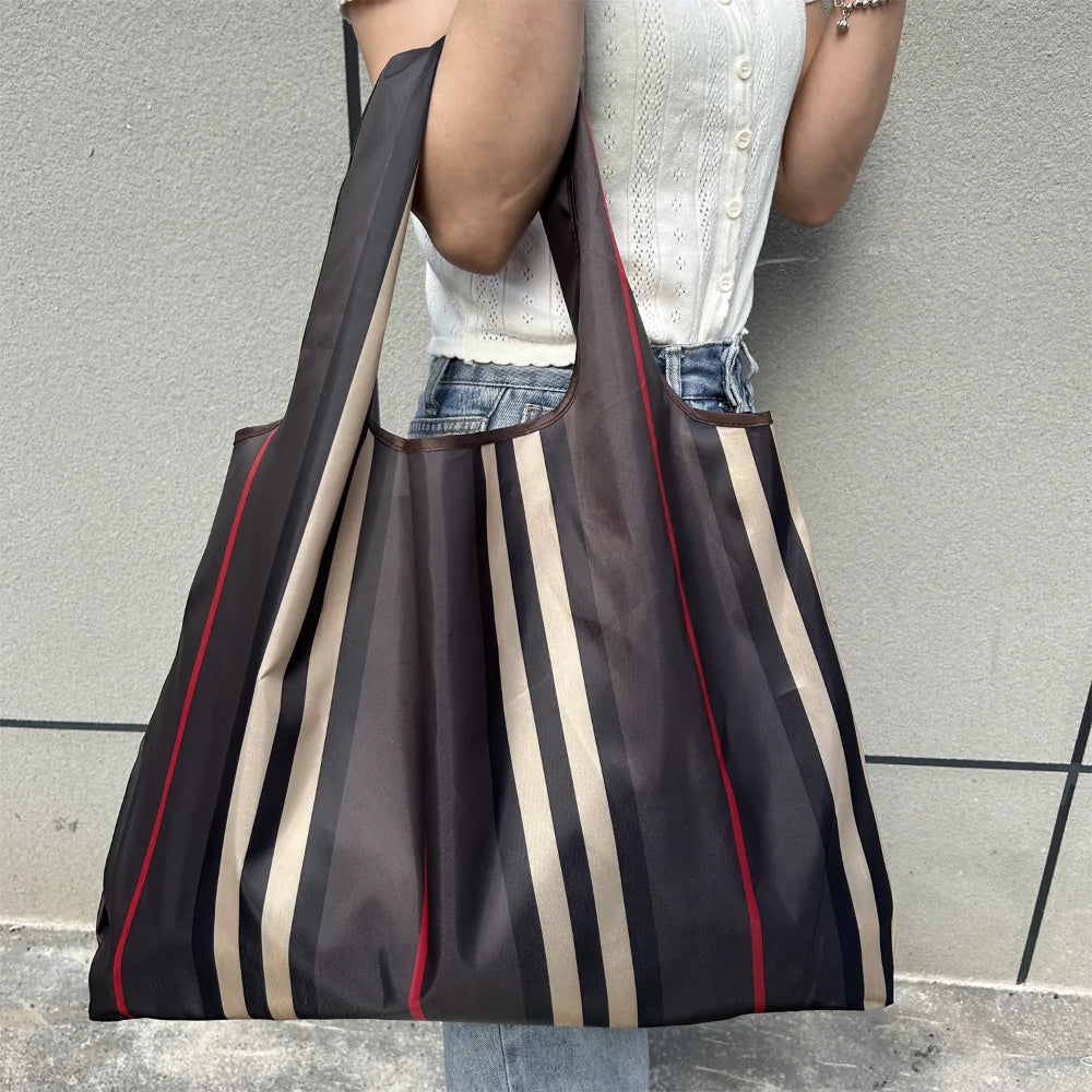 A person is holding a dark brown and beige striped foldable oxford grocery tote bag against a gray wall.