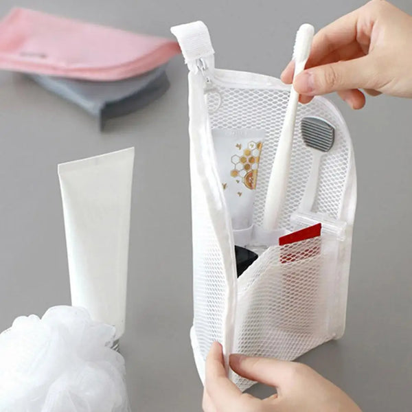 A hand is placing a white toothbrush and a small cosmetic item into a clear mesh travel bag.