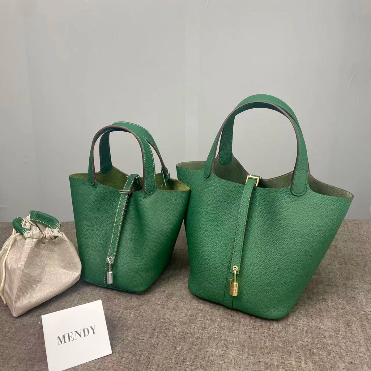A dark green genuine leather bucket bag with a strap is displayed on a gray surface next to a name tag.
