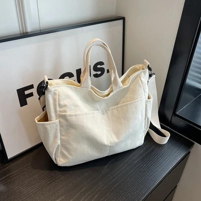 A cream corduroy multi-pocket tote bag with light vertical ribbing is displayed on a dark surface.