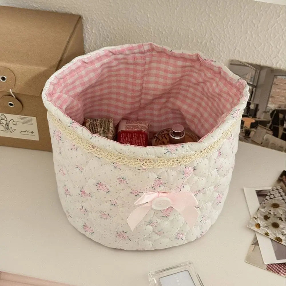 Cesta de algodón acolchada rosa y blanca con forma de corazón, llena de objetos pequeños sobre una superficie blanca.