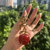 A hand holds a gold-toned crystal fruit charm keychain with a red, glittery apple.