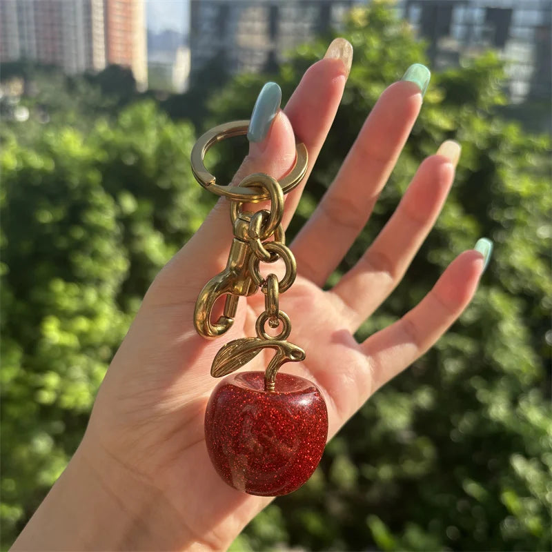 A hand holds a gold-toned crystal fruit charm keychain with a red, glittery apple.