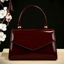 A glossy burgundy evening handbag with a leather strap and gold hardware resting on a light wooden surface.