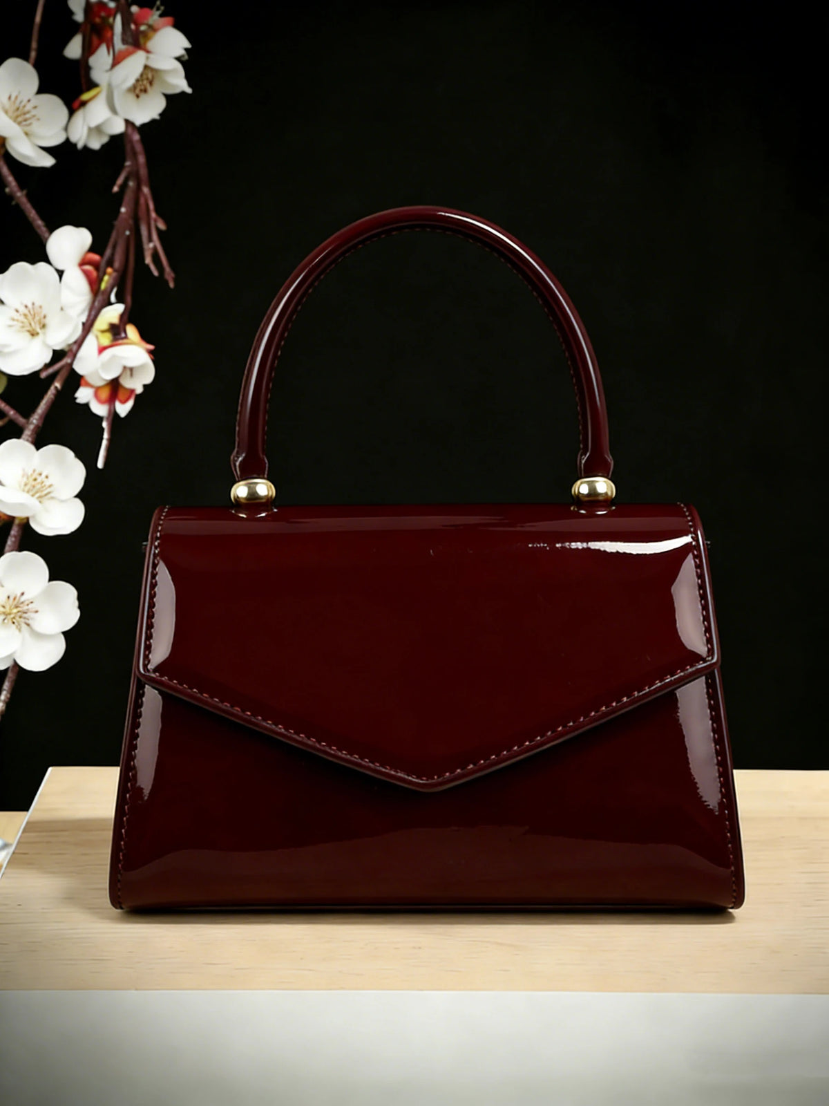 A glossy burgundy evening handbag with a leather strap and gold hardware resting on a light wooden surface.