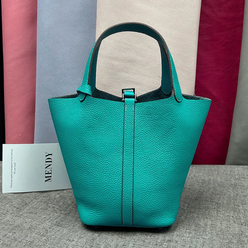 A teal leather bucket bag with a dark green strap is displayed against a backdrop of various colored fabric.