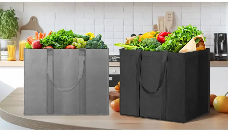 A set of three reusable black foldable grocery tote bags filled with fresh vegetables on a kitchen counter.