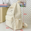 A beige nylon backpack with a decorative embroidery detail is shown next to a ruler for scale.