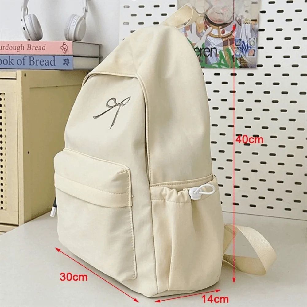 A beige nylon backpack with a decorative embroidery detail is shown next to a ruler for scale.