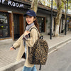 A woman wearing a beige coat and jeans walks past a building with tree-lined pavement, carrying a leopard print canvas backpa