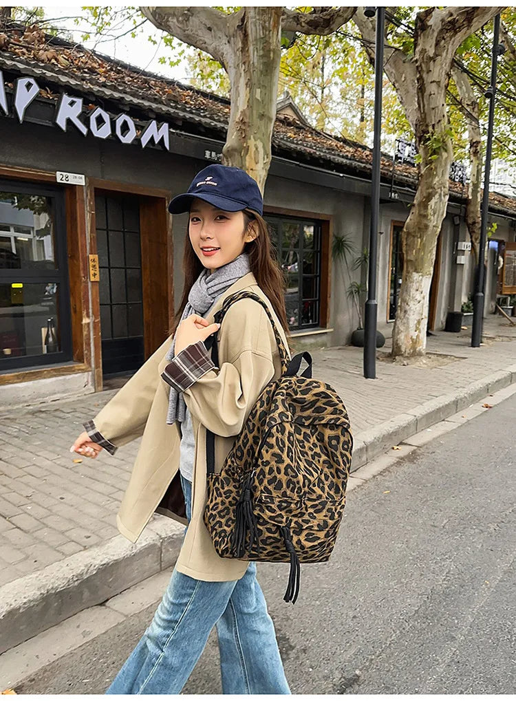 A woman wearing a beige coat and jeans walks past a building with tree-lined pavement, carrying a leopard print canvas backpa