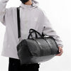 A person wearing a light gray hooded jacket holds a black leather-trimmed travel duffle bag.