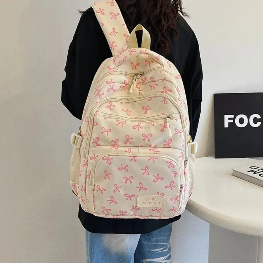 A light pink and cream nylon backpack with a repeating bow print, worn by a person.