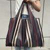 A dark brown and beige striped foldable oxford grocery tote bag is being held by a person against a light gray background.