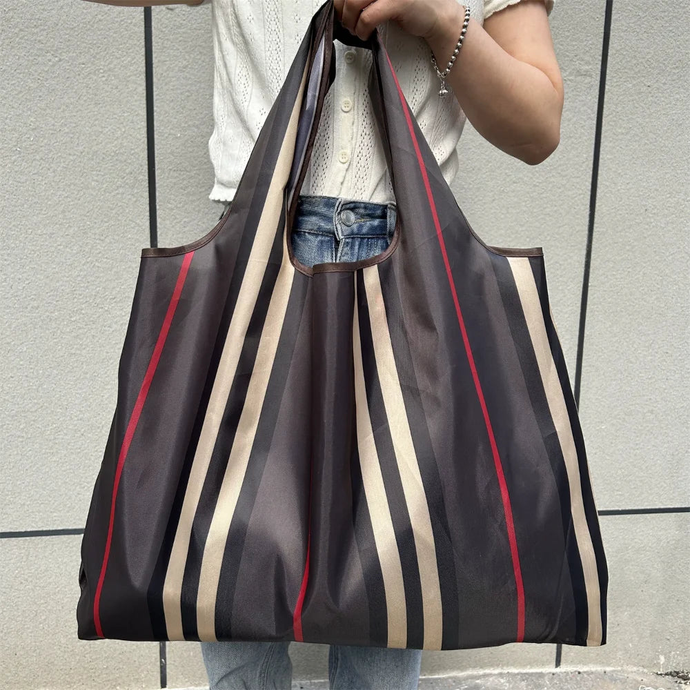 A dark brown and beige striped foldable oxford grocery tote bag is being held by a person against a light gray background.