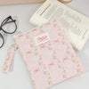 A pink floral plaid cotton tablet pouch with a secure closure resting on a white surface next to a book and a mug.