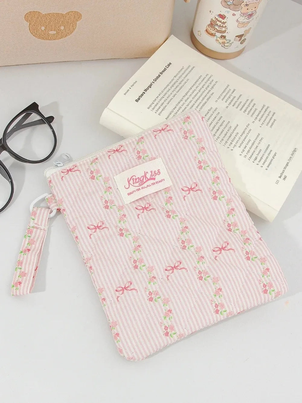 A pink floral plaid cotton tablet pouch with a secure closure resting on a white surface next to a book and a mug.