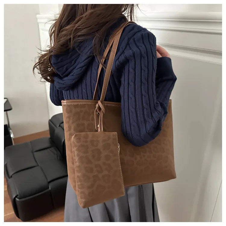 A woman holds a large brown leopard print tote bag with a leather strap against a white wall.