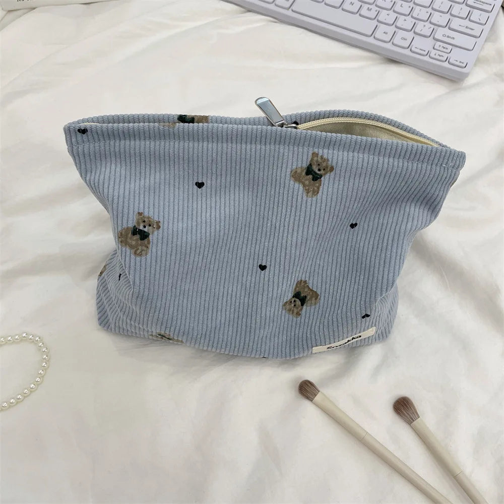 A light blue corduroy clutch bag with small brown bear embroidery, lying on a white surface.
