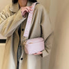 A woman wearing a beige trench coat holds a light pink vegan PU crossbody bag with a patterned strap.