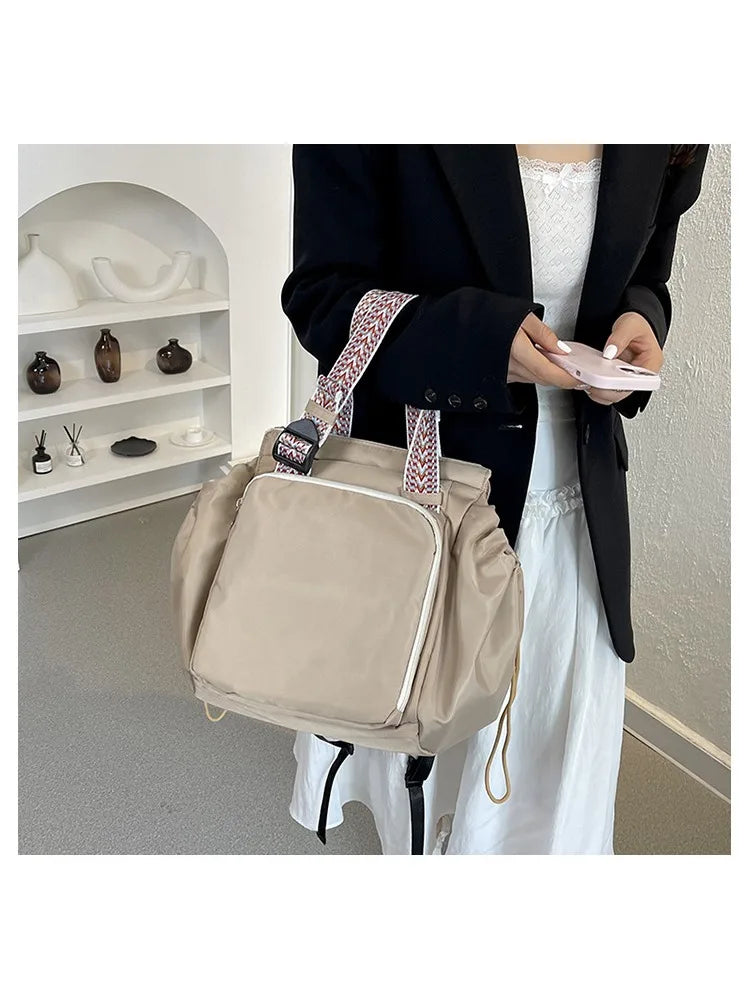 A beige nylon shoulder bag with a patterned strap is held by a person in a black blazer.